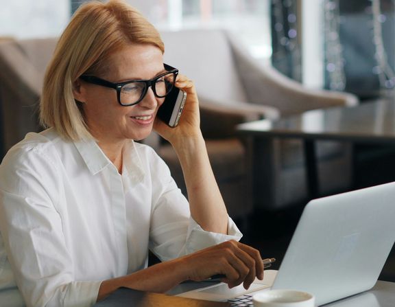 Frau mit blonden Haaren und Brille sitzt im Büro, arbeitet am Laptop, telefoniert und lächelt dabei