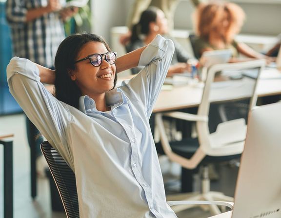 Frau sitzt im Büro am Schreibtisch und sieht sehr zufrieden ist. Im Hintergrund sind noch weitere Personen an Schreibtischen zu sehen